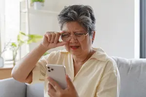 An older female hartland eye care patient is sitting on a couch while holding a cell phone in her hand and adjusting her eyeglasses.