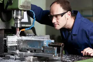 A man wearing safety glasses and a blue work shirt operates a metalworking machine in a workshop.