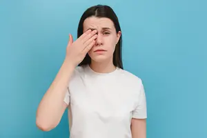 A woman wearing a white shirt is holding her eye with her left hand and her head is slightly tilted to the left. She is probably posing for a photograph. She has a serious expression on her face. She is probably looking at something with a sad expression. The background is a solid blue color. The light is focused on her face.