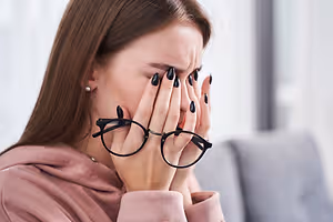 Closeup of a young woman sitting on a couch rubbing her eyes while holding her glasses