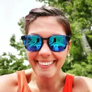 woman with short hair wearing sunglasses with a reflection of a building and trees in them smiling for a selfie