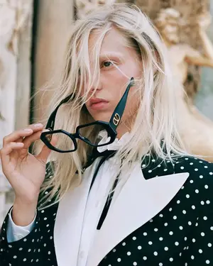 Blonde woman wearing a black and white polka dot suit jacket with a white shirt and black glasses with a gold logo on the corner