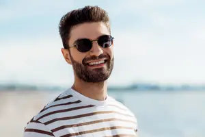 A smiling man with sunglasses and a striped shirt standing by the beach