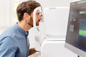 A man wearing a blue shirt is sitting in front of a monitor while looking at the machine, which is probably an eye exam machine, and he is probably doing an eye exam.