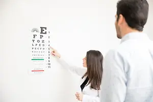 A woman in a white lab coat points to an eye chart on a white wall, with a man in a white shirt looking at her from behind.