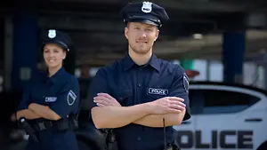 Two police officers in uniform, one with folded arms, standing beside a police car in a garage.