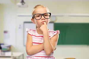 A little girl wearing glasses and a pink striped dress is standing in a classroom and posing for a photo.