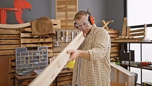 A person wearing goggles and headphones is holding a wooden plank in a workshop with various items like tools and chairs.