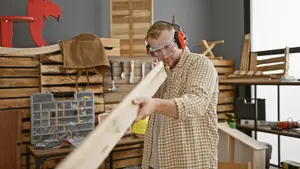 A person wearing goggles and headphones is holding a wooden plank in a workshop with various items like tools and chairs.