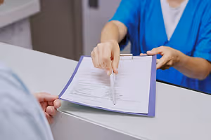 A woman in a blue uniform is filling out a form with a pen while a man is looking at her in a hospital setting.