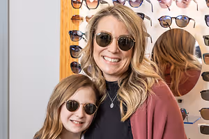 A woman and a young girl are smiling and posing for a photo in front of a wall with sunglasses on display.