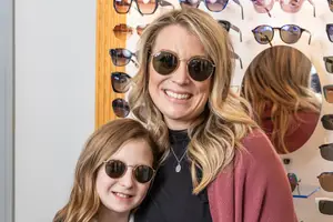 A woman and a young girl are smiling and posing for a photo in front of a wall with sunglasses on display.