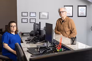 Two people, one sitting and one standing, pose in front of a desk with computers and various office supplies.