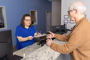 An elderly man in a brown sweater is speaking with a woman wearing a blue shirt and glasses at a reception desk in a medical office.