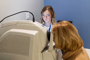 A woman wearing glasses is using an ophthalmoscope to look into a patient's eye in a medical room.