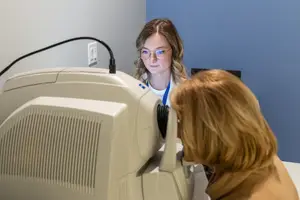 A woman wearing glasses is using an ophthalmoscope to look into a patient's eye in a medical room.
