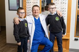 A man in a white coat and blue scrubs stands with two young boys in front of a mirror in a room with wooden flooring and a white wall with a framed picture and a wall of eyeglasses, posing for a photo