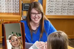 A smiling woman wearing glasses adjusts a young girl's glasses in front of a mirror in an optician's shop.