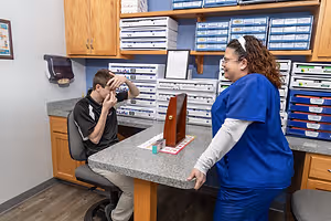 A woman wearing a blue uniform is standing in front of a desk, while a man wearing a black and white shirt is sitting in a chair and holding his head.