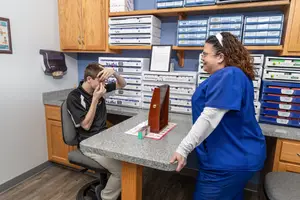 A woman wearing a blue uniform is standing in front of a desk, while a man wearing a black and white shirt is sitting in a chair and holding his head.