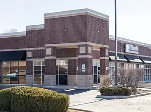An exterior view of a storefront featuring a brick facade, large glass windows, and a green sign on the left side.