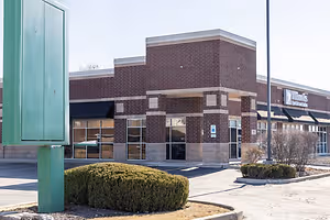 An exterior view of a storefront featuring a brick facade, large glass windows, and a green sign on the left side.