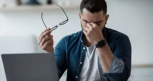 Man holding eyeglasses and pinching the bridge of his nose beside a laptop