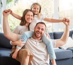 A joyful family of three, with a child sitting on a man's shoulders, all smiling in a bright living room.