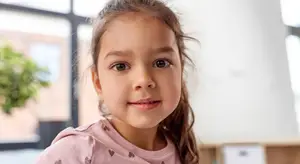 A young girl with brown hair and wearing a pink shirt with heart designs is smiling and looking at the camera. She is standing in front of a glass wall that shows a blurry view of a plant and a building.
