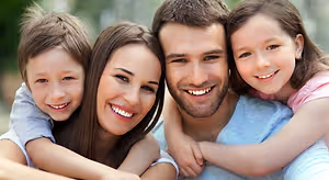 A family of four, including a man, a woman, and two children, are smiling and posing for a photograph in a park, with a blurry background of trees and plants