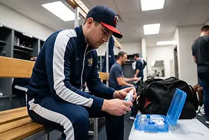 A man sitting on a bench in a locker room opening a bottle of lotion.