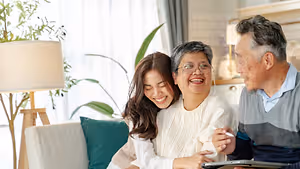 A family of three, an elderly man and woman and a young woman, sitting on a couch in the living room, smiling and looking at something, possibly a book, in front of them. Behind them is a plant, a lamp, and a cabinet with a lamp on top. The lamp is casting a warm glow on the room.