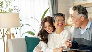 A family of three, an elderly man and woman and a young woman, sitting on a couch in the living room, smiling and looking at something, possibly a book, in front of them. Behind them is a plant, a lamp, and a cabinet with a lamp on top. The lamp is casting a warm glow on the room.