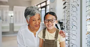An older woman with gray hair and a younger girl with black hair wearing glasses are standing and smiling in front of a glass display case in a room with a white wall and a glass door.