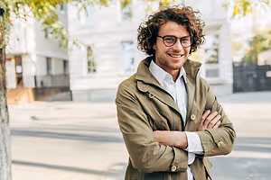 A smiling man with curly hair stands on a street, wearing a light green jacket, glasses, and a white shirt, with his arms crossed and a tree and buildings in the background.