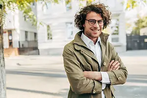 A smiling man with curly hair stands on a street, wearing a light green jacket, glasses, and a white shirt, with his arms crossed and a tree and buildings in the background.