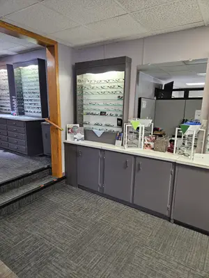 A room with gray furniture, a wooden column, and a staircase, filled with various items including eyeglasses, mirrors, cabinets, and a desk, creating a space that combines functionality and style, possibly an office or a boutique.