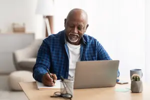 A bald man with a beard wearing a blue shirt sits at a desk with a laptop and writes in a notebook.