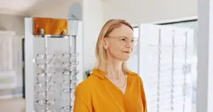An older woman with blonde hair wearing glasses stands in front of a display of eyeglasses in a shop.