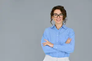 A woman is standing with her arms folded, smiling, and looking at the camera, wearing a blue striped shirt and glasses.