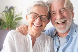 Elderly couple laughing and posing for a photo inside a home with a potted plant in the background
