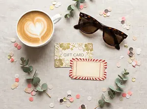 A coffee cup with a heart design in the foam and a gift card on a table with confetti and sunglasses.