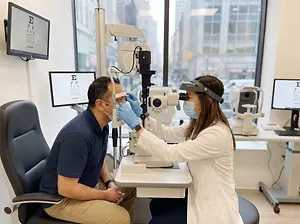 An optometrist is examining a patient's eyes in an office with a large window overlooking a city street.