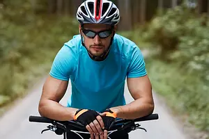 A man in a blue t-shirt, black pants, and a helmet rides a bicycle on a dirt road surrounded by green grass and trees.