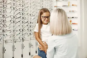A girl in glasses is standing with a woman who is standing behind her and holding her waist inside a shop with shelves with eyeglasses on them.