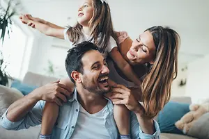 A man is sitting on the couch with a woman and a child on his shoulders, smiling at the camera.