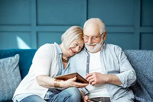 An elderly couple sitting on a couch, looking at a tablet with smiles on their faces