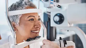 A smiling woman with gray hair is having her eye examined by an optometrist using a slit lamp.