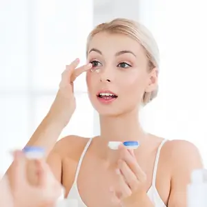 A woman wearing a white tank top is looking at herself in a mirror while applying contact lenses.