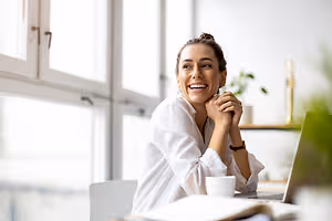 A smiling woman sitting at a desk with a laptop, cup, and books, wearing a white shirt and a bracelet, in a room with glass windows and a plant in the corner.